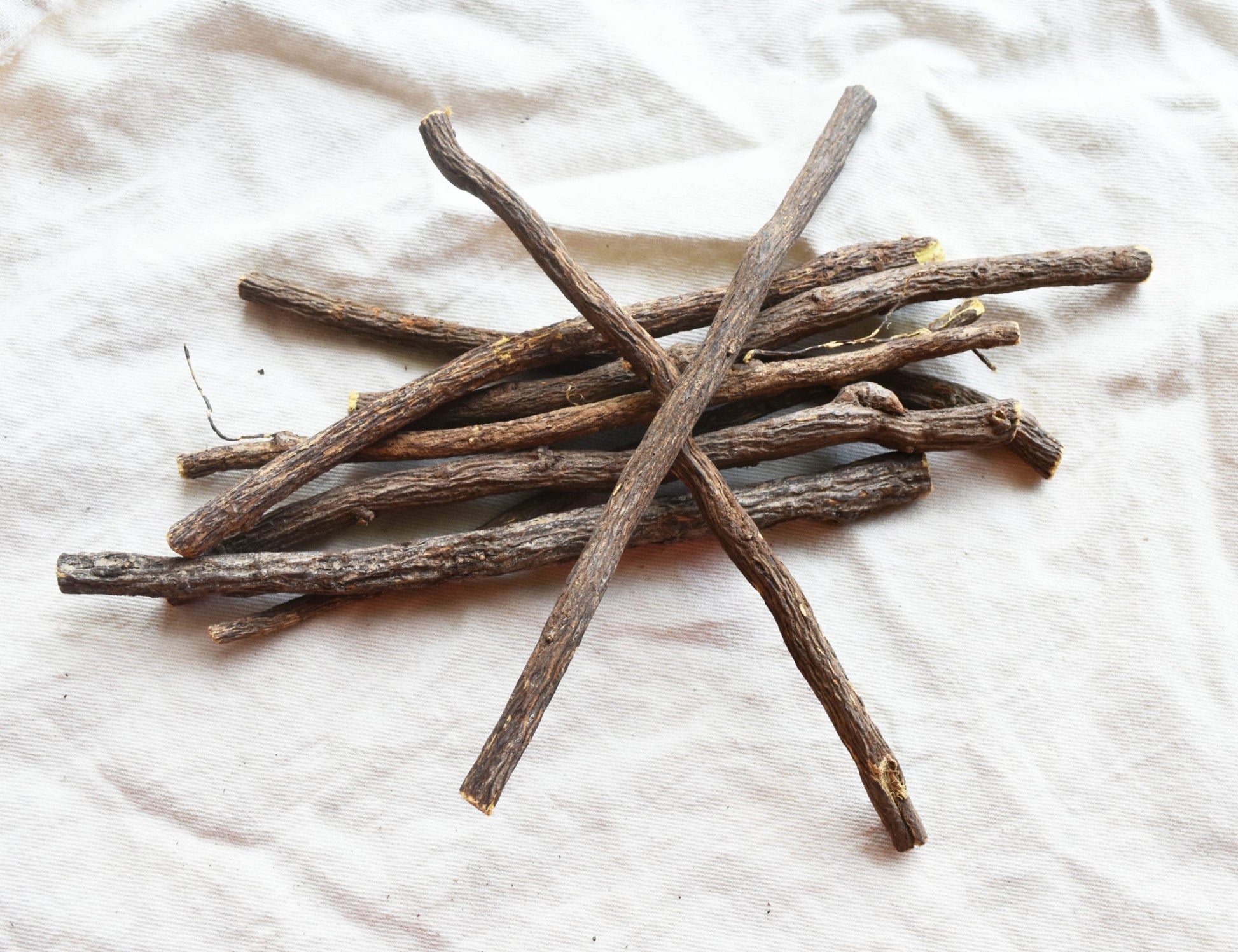 The image presents a close-up, overhead view of a small pile of dried licorice root sticks arranged haphazardly on a textured, off-white fabric backdrop, likely canvas or linen. The licorice pieces vary in length, ranging from approximately 3 to 6 inches, and are roughly cylindrical in shape, though naturally irregular. Their color is a muted, earthy brown with subtle variations in tone, suggesting different degrees of drying and processing. The surface of the licorice root is visibly textured, displaying a slightly rough, fibrous appearance with occasional darker patches and small imperfections. Several sticks are laid parallel to each other, while others cross over them at angles, creating a somewhat chaotic yet natural arrangement. The fabric beneath the licorice root is wrinkled and has a visible weave, adding to the organic feel of the image. The lighting appears soft and diffused, casting gentle shadows that highlight the texture of both the licorice and the fabric. The overall composition is simple and focuses entirely on the natural form and texture of the licorice root, suggesting a focus on its raw, unprocessed state. The image evokes a sense of natural remedies, herbalism, or traditional ingredients.